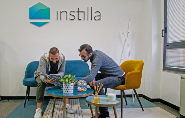 Due businessman seduti nel salottino di un'azienda, mentre guardano una rivista. Alle spalle il logo aziendale: Instilla.